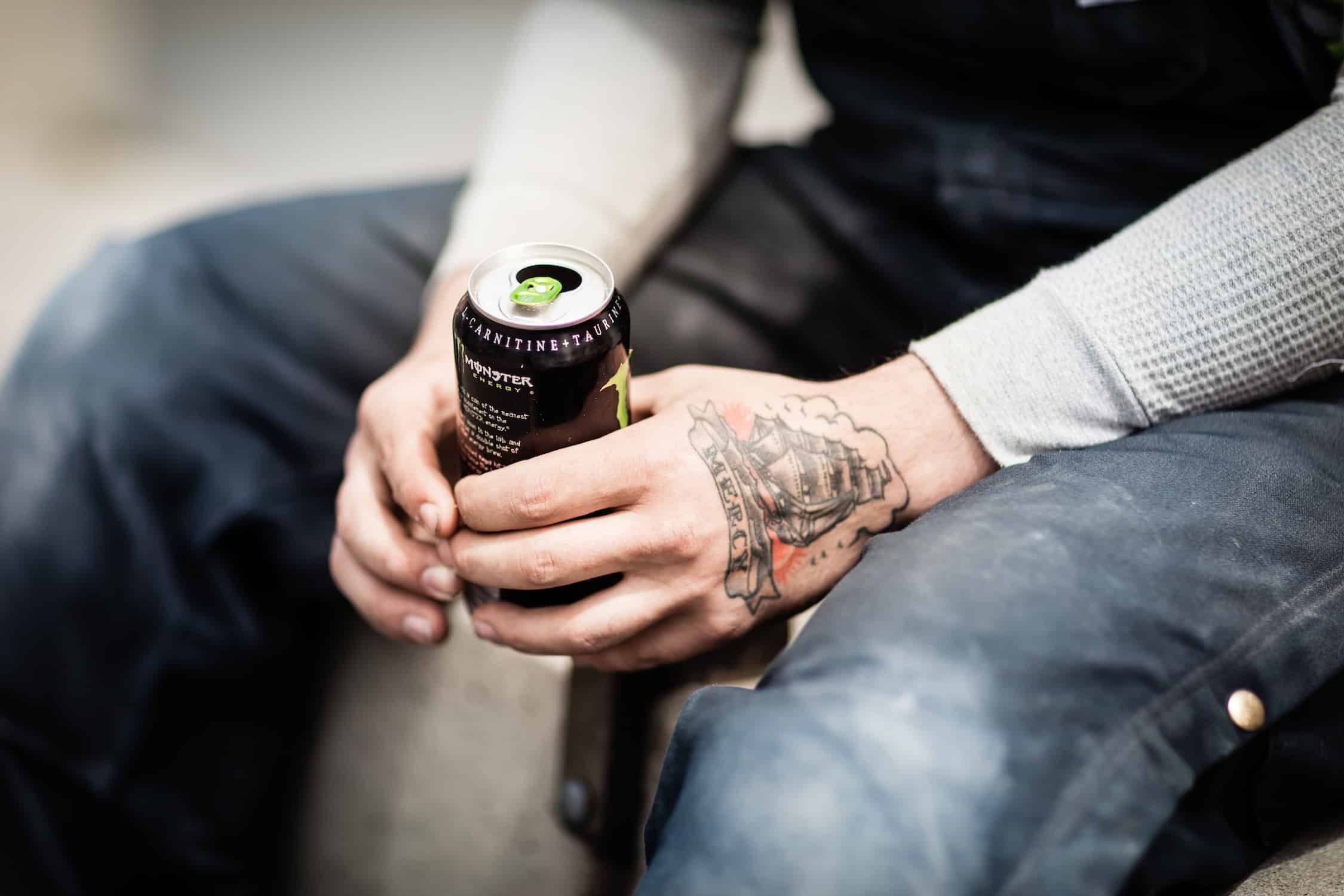 Two people holding a beverage can together, outdoor casual setting with visible tattoo on arm.