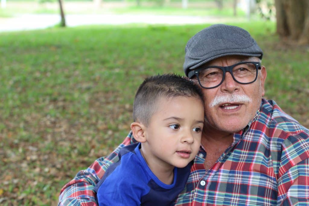 Elderly man and young boy smiling together outdoors in casual plaid shirts near grass and trees.