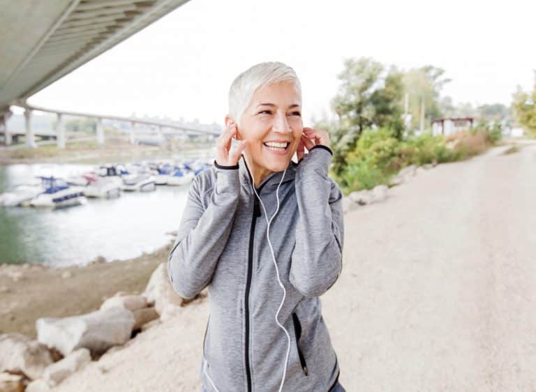 Older woman with gray hair smiling outdoors near a bridge and parking lot.