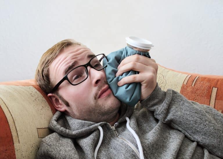 Man wearing glasses lying on couch, holding dental mirror for self-examination indoors.